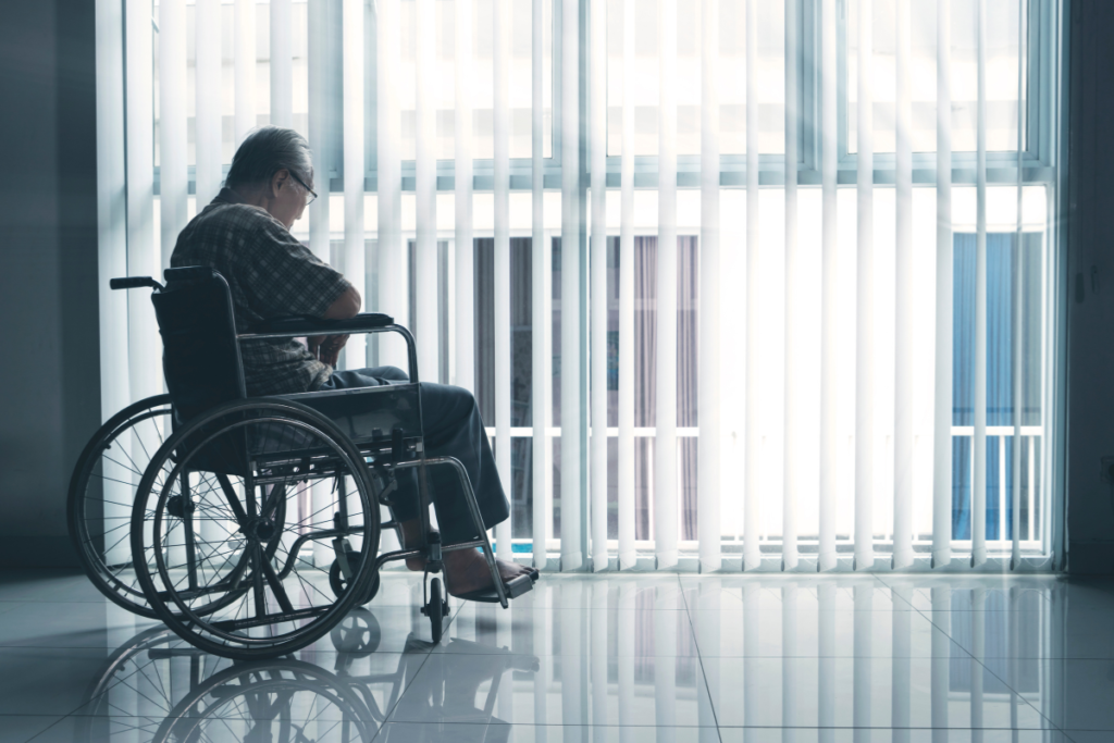 An elderly man sitting in a wheelchair looking out of a window.