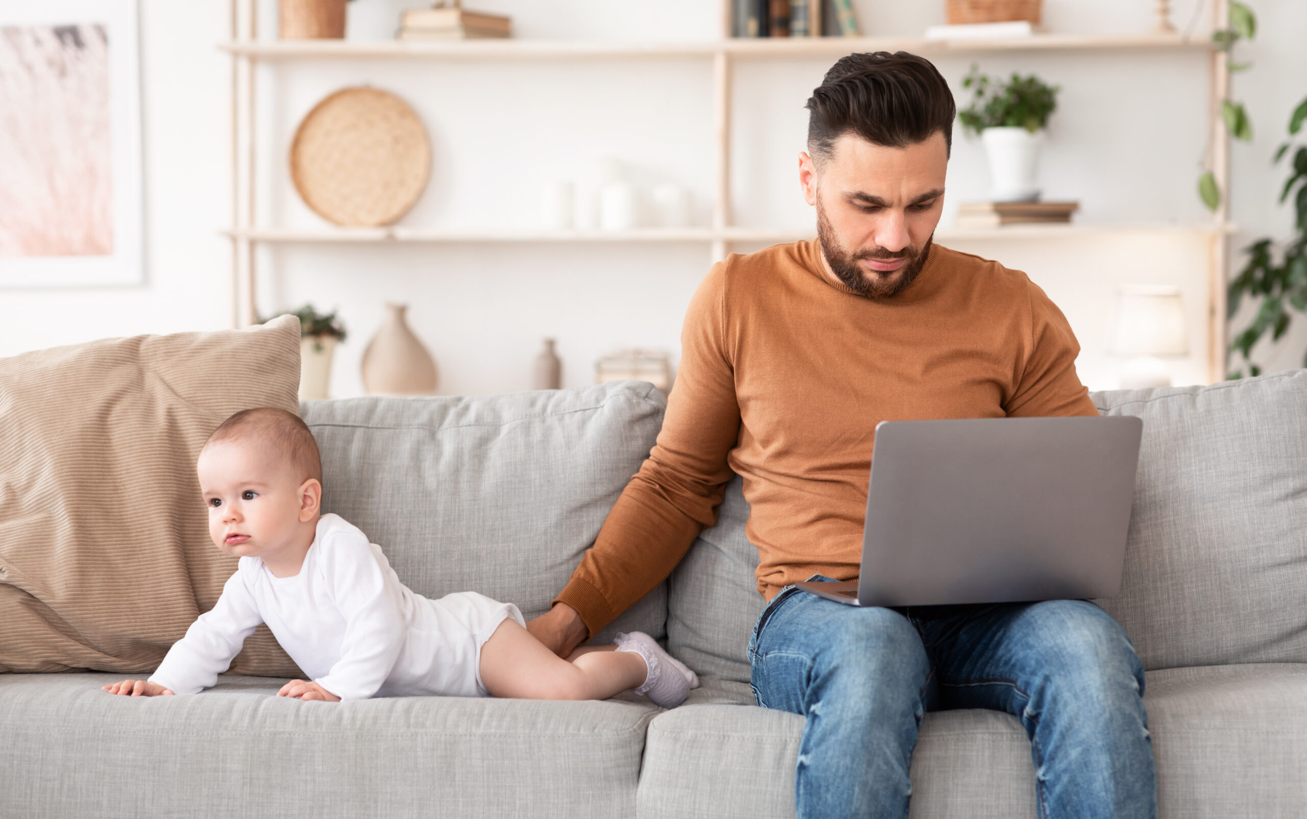 Young Father With Baby Using Laptop Browsing Internet Working Online Sitting On Sofa At Home
